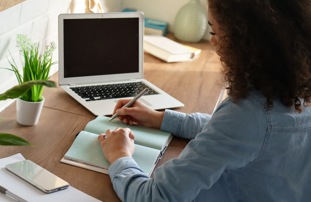 female office worker sitting in front of laptop writing in a notebook